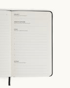 Open spread of the black Matine Daily Planner showing the “Project,” “Today’s Outcome,” “Steps,” and “Extras” page with numbered lines, check circles, and ribbon bookmark visible.