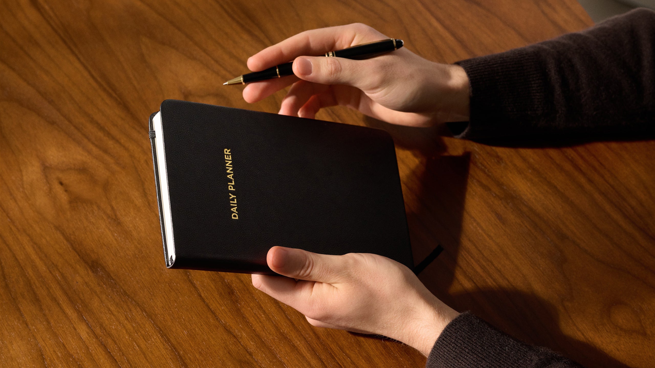 Hands holding a closed black daily planner with gold title on a wooden table.
