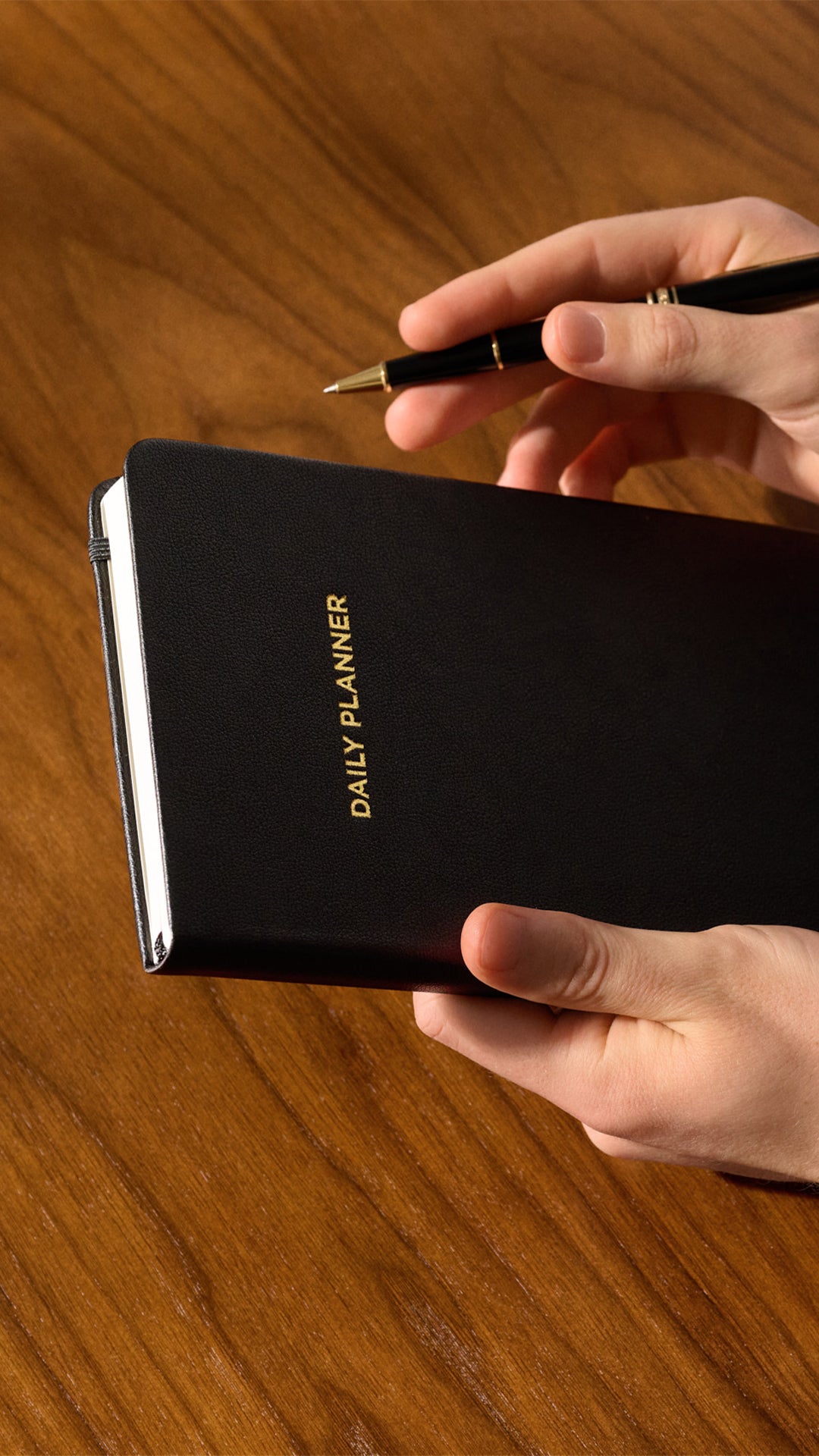Hands holding a closed black daily planner with gold title on a wooden table.