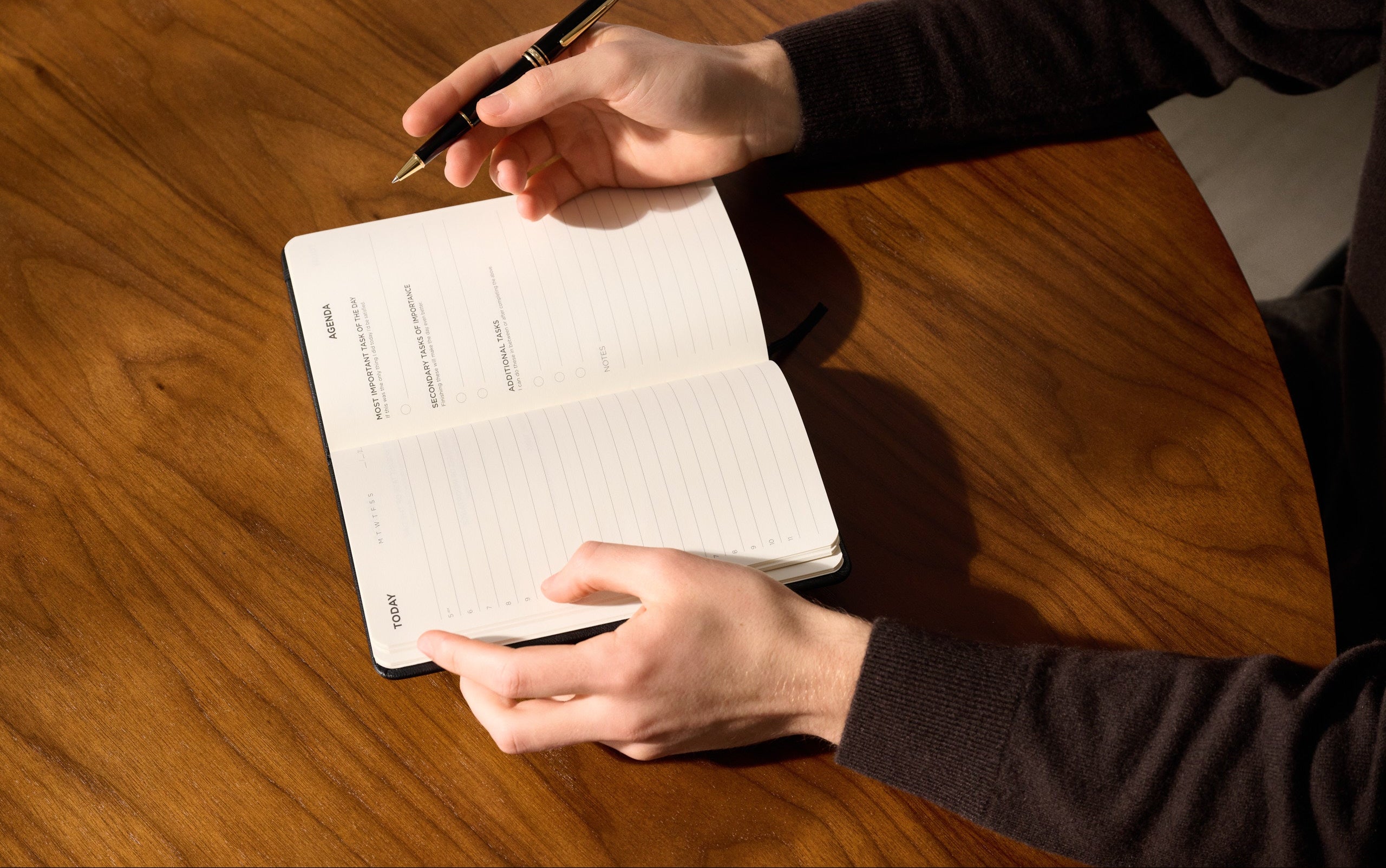 Person schreibt mit einem Stift in einen offenen Daily Planner auf einem Holztisch.