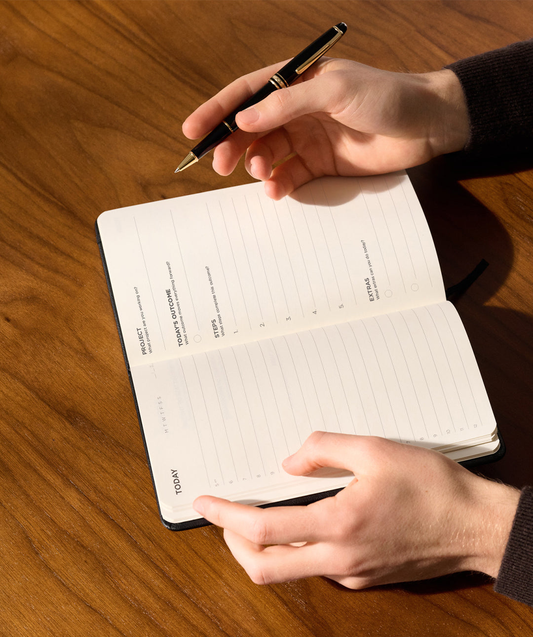 Hands holding an open black Matine Daily Planner showing the “Today,” “Project,” “Today’s Outcome,” and “Steps” pages with a black pen above a wooden desk.