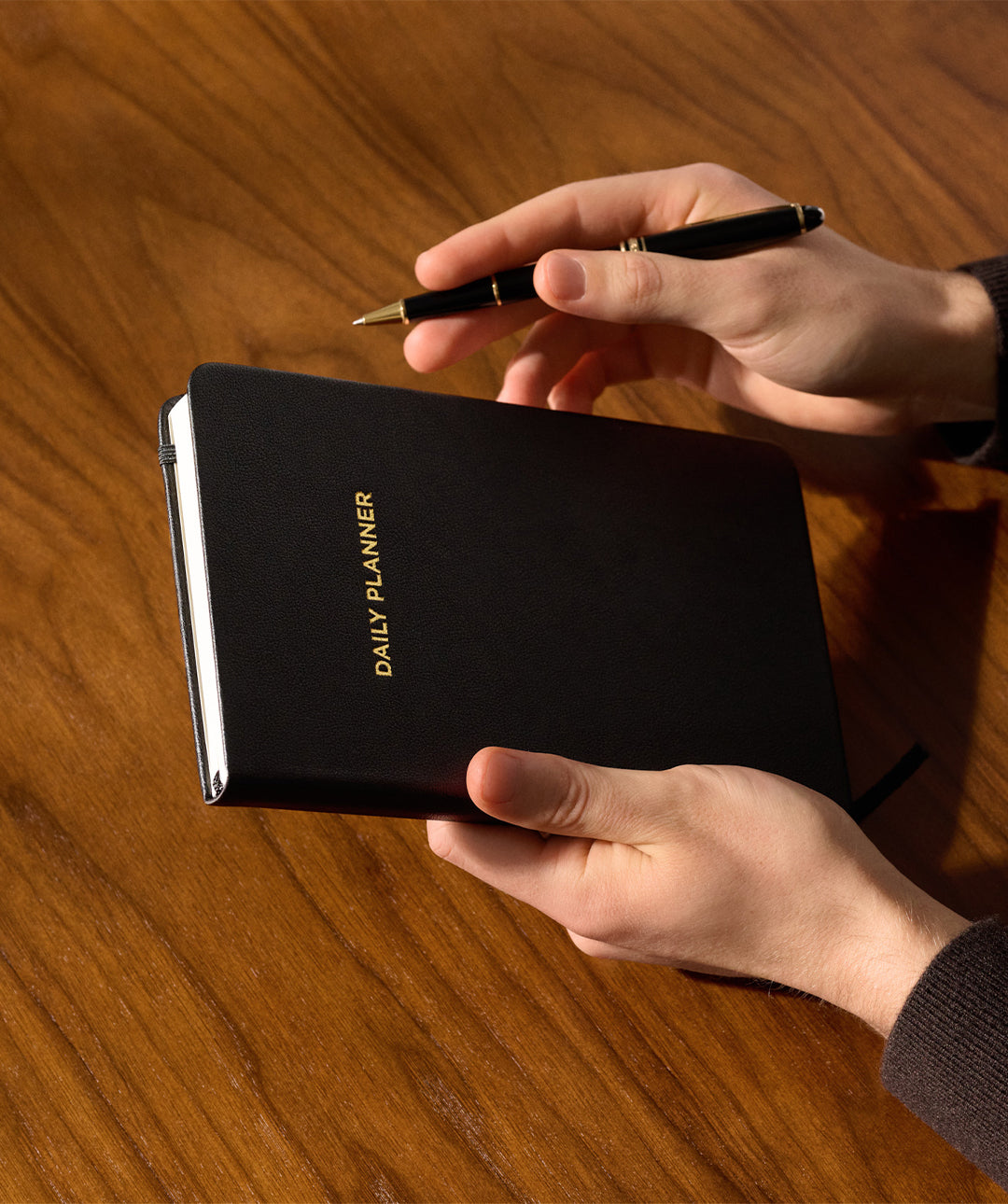 Hands holding the black Matine Daily Planner with gold “DAILY PLANNER” embossing and a black pen above a wooden desk.